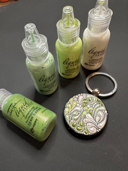 Fünf Flaschen mit grüner und weißer Farbe neben einem dekorativen Schlüsselanhänger.