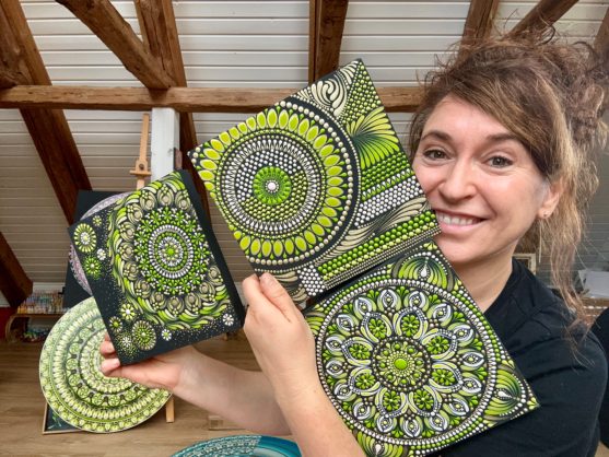 Frau lächelt und hält mehrere bunte Mandalas in Grün- und Grautönen hoch.